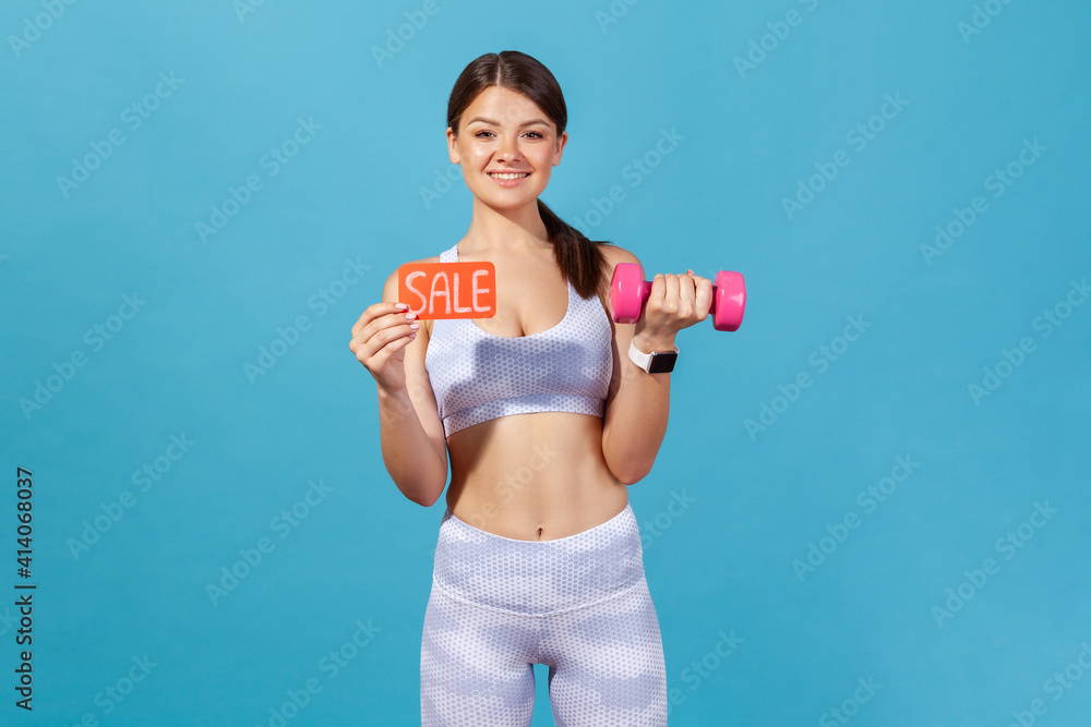 Smiling happy woman in white sportswear holding in hands dumbbell and sale card, buying gym membership and private lessons with coach. Indoor studio shot isolated on blue background Smiling happy woman in white sportswear holding in hands dumbbell and sale card, buying gym membership and private lessons with coach. Indoor studio shot isolated on blue background