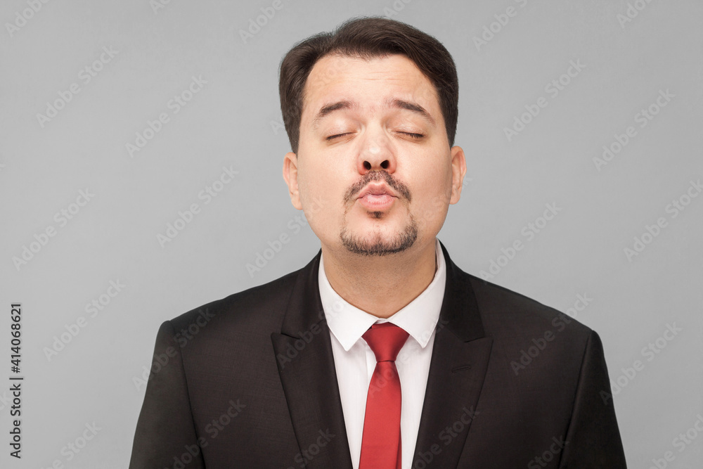 Concept of love. Amorous man kissing an imaginary girl getting ready for a date. indoor studio shot, isolated on gray background. businessman with black suit, red tie and mustache looking at camera. Concept of love. Amorous man kissing an imaginary girl getting ready for a date. indoor studio shot, isolated on gray background. businessman with black suit, red tie and mustache looking at camera.