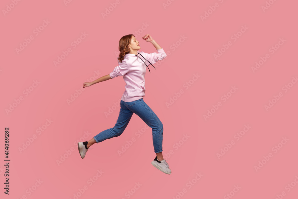 Full length side view of young female in hoodie and jeans running in air, hurrying for discounts, empty copy space for advertising. Indoor studio shot isolated on pink background