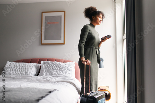 Woman With Suitcase Checking Out Of Boutique Hotel Waiting For Taxi Ordered On Mobile Phone App