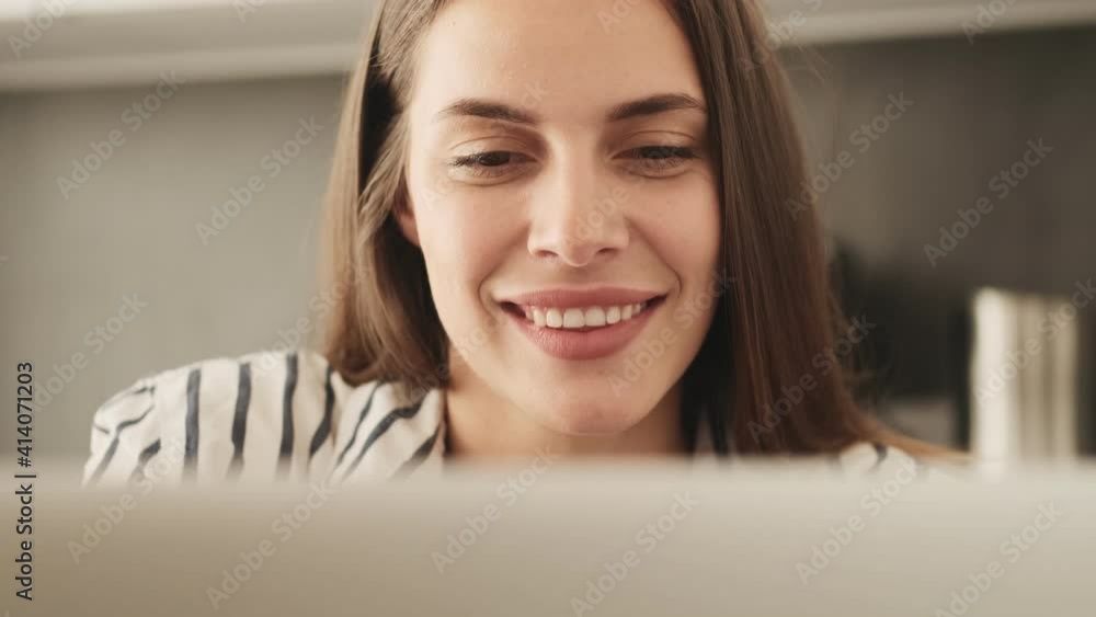 A close-up view of a smiling happy young woman is using her laptop computer while sitting in the kitchen at home A close-up view of a smiling happy young woman is using her laptop computer while sitting in the kitchen at home