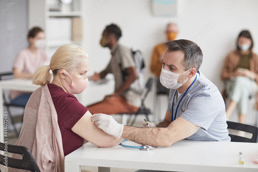 Portrait of mature male doctor vaccinating senior patient in vaccination center or clinic, both wearing masks, copy space