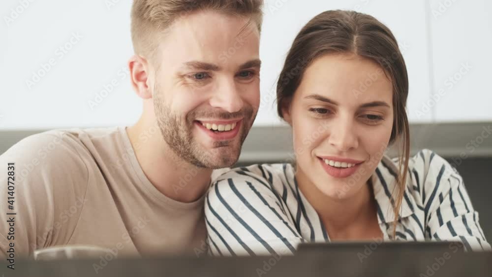 A pleased young couple man and woman are sitting and watching video in the kitchen at home A pleased young couple man and woman are sitting and watching video in the kitchen at home