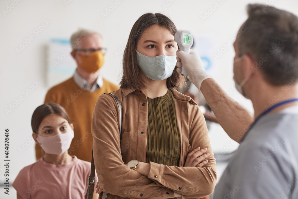 Shot of unrecognizable doctor checking temperature of young woman wearing mask and waiting in line at clinic or hospital, copy space