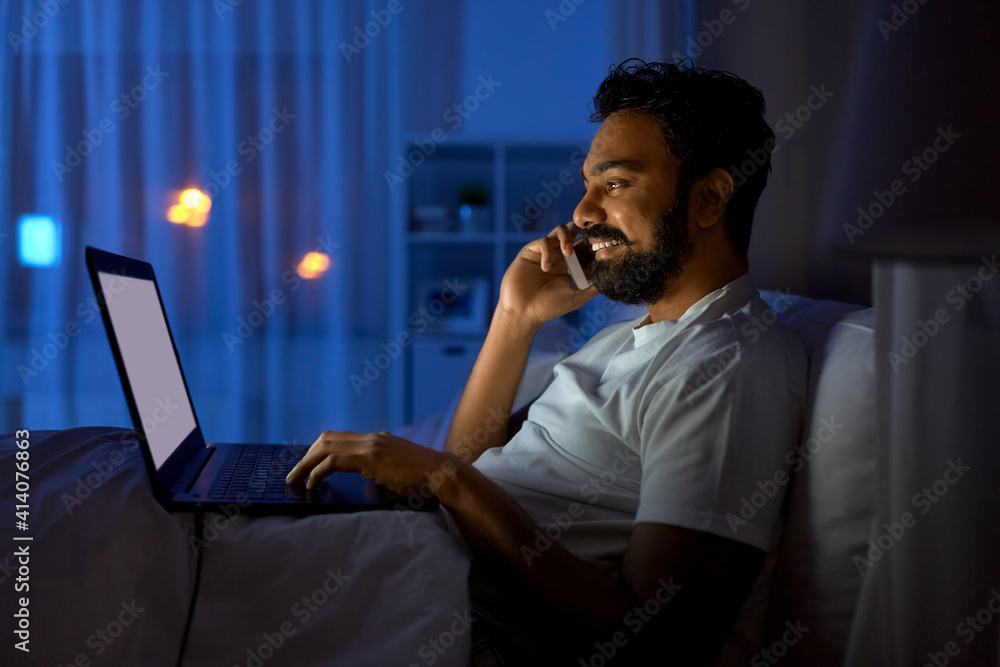 technology, remote job and people concept - happy smiling young indian man with laptop computer calling on smartphone in bed at home at night technology, remote job and people concept - happy smiling young indian man with laptop computer calling on smartphone in bed at home at night