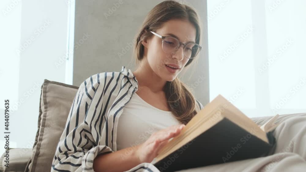 A concentrated young woman wearing glasses is reading book sitting on the sofa at home A concentrated young woman wearing glasses is reading book sitting on the sofa at home