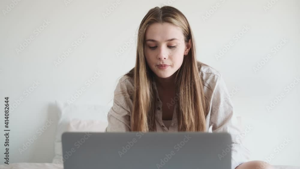 A positive woman is talking with the video connection using her laptop computer while sitting in the bed at the white bedroom at home A positive woman is talking with the video connection using her laptop computer while sitting in the bed at the white bedroom at home