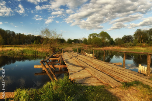 Wooden footbridge from old boards and reflection of the sky with clouds in the water of a calm lake. Morning rural landscape on the river. © Ann Stryzhekin