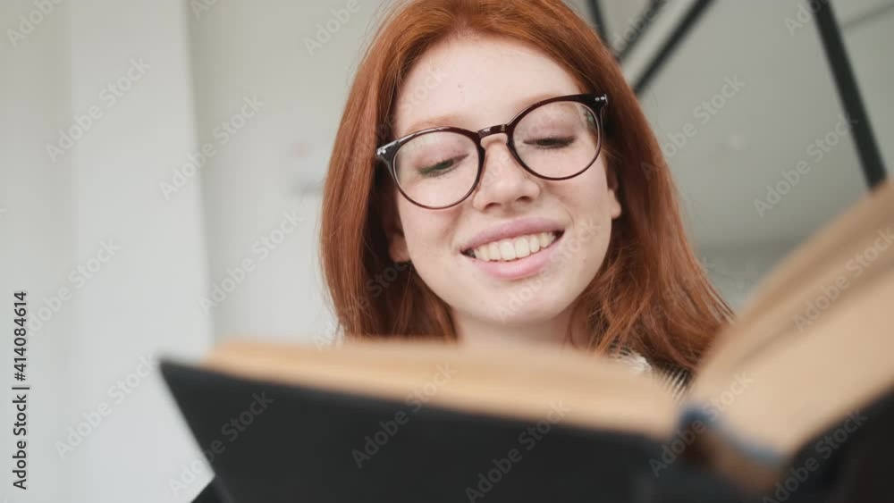 A nice smiling redhead woman wearing glasses is reading a book sitting in the apartments A nice smiling redhead woman wearing glasses is reading a book sitting in the apartments