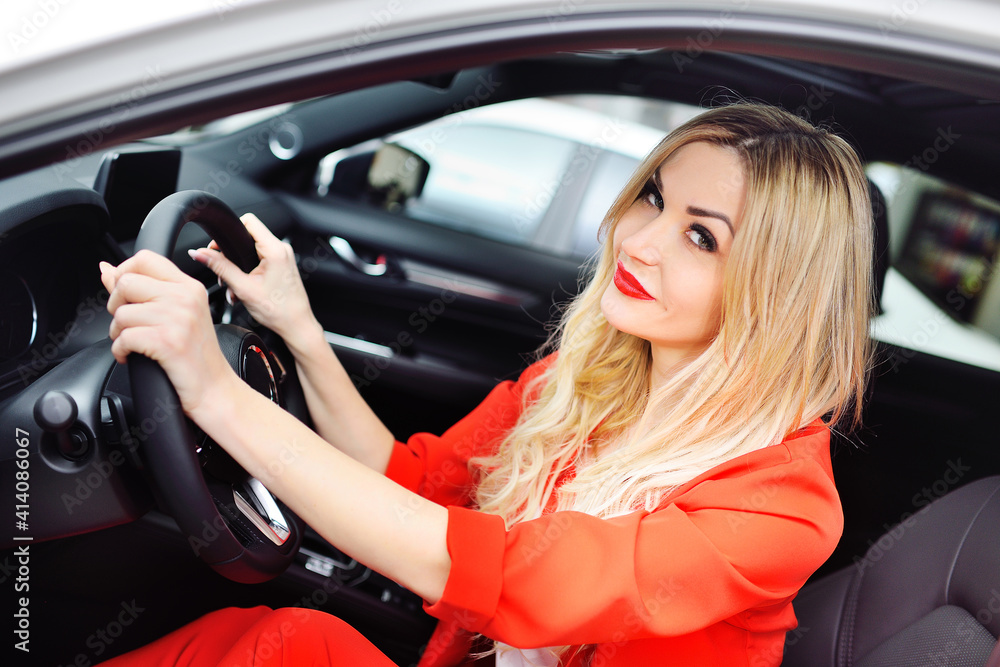 a young attractive blonde woman in a red suit sits behind the wheel of a car and smiles.