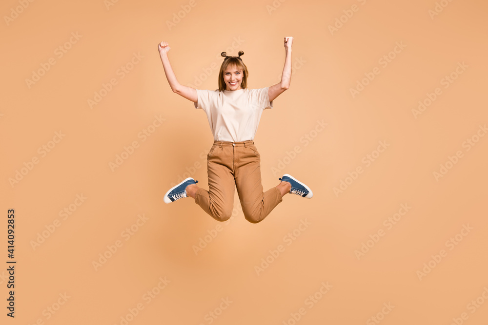 Full length photo of girl jump raise fists wear t-shirt trousers footwear isolated beige color background