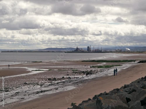 People walk along a dirty looking beach with sea fall defense and industrial landscape in the background photo