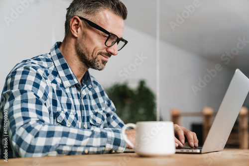 Pleased unshaven man in eyeglasses working with laptop in office