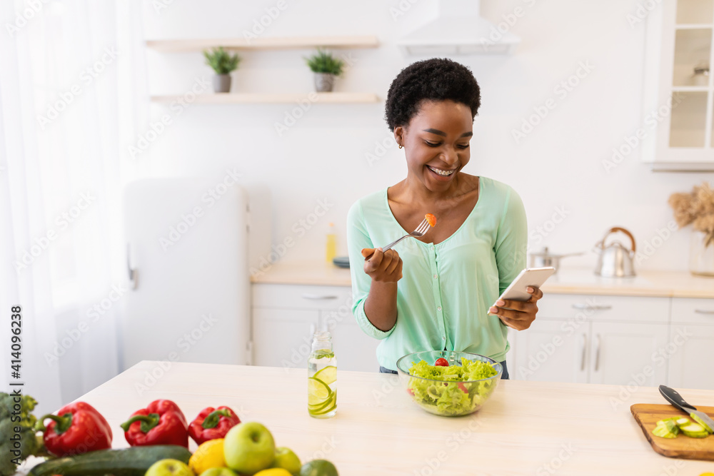 Black Woman Using Smartphone Eating Vegetable Salad In Modern Kitchen Black Woman Using Smartphone Eating Vegetable Salad In Modern Kitchen
