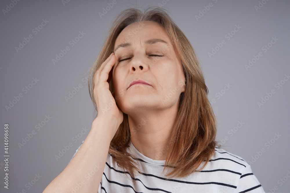 Fototapeta premium senior woman in a striped shirt is holding her head, suffering from pain. Isolate on gray background