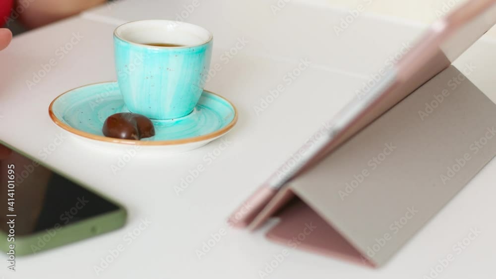 Drink coffee in the morning. Woman's hand takes a cup of hot drink from the table in the kitchen while viewing the tablet. Many gadgets on the table Drink coffee in the morning. Woman's hand takes a cup of hot drink from the table in the kitchen while viewing the tablet. Many gadgets on the table