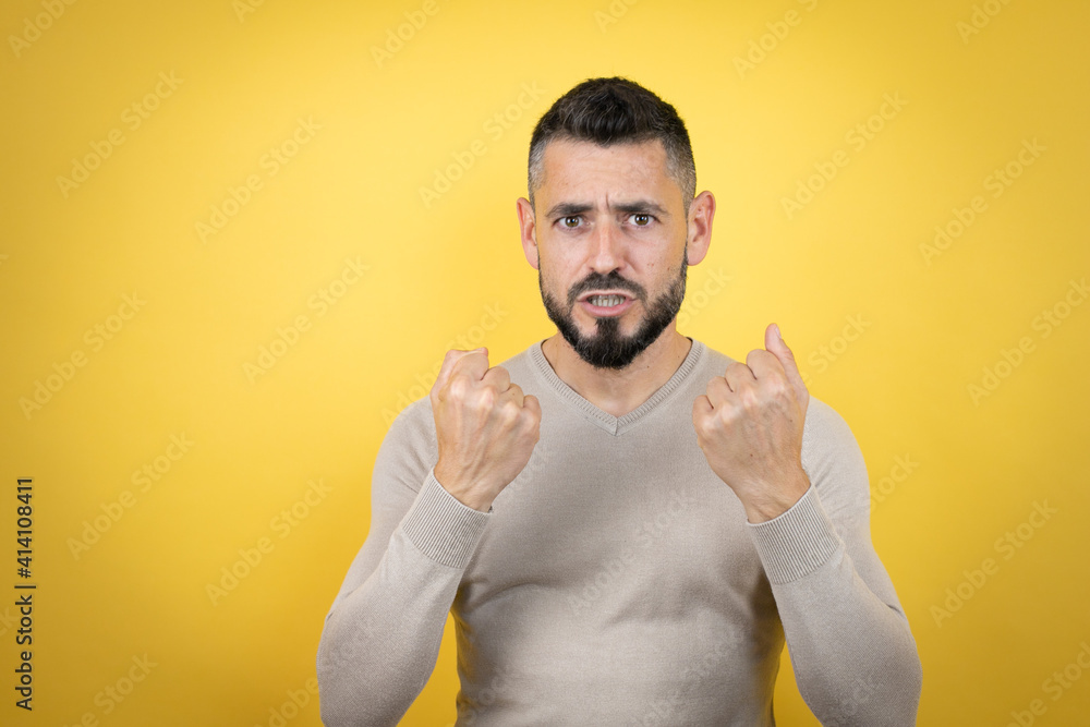 Handsome man with beard wearing sweater over yellow background very happy and excited making winner gesture with raised arms, smiling and screaming for success. Handsome man with beard wearing sweater over yellow background very happy and excited making winner gesture with raised arms, smiling and screaming for success.