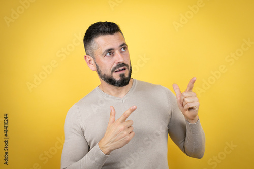 Handsome man with beard wearing sweater over yellow background surprised and pointing her fingers side