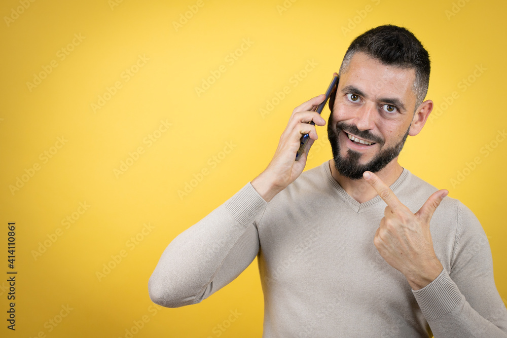 Handsome man with beard wearing sweater over yellow background having conversation talking on the smartphone smiling happy pointing with hand and finger Handsome man with beard wearing sweater over yellow background having conversation talking on the smartphone smiling happy pointing with hand and finger