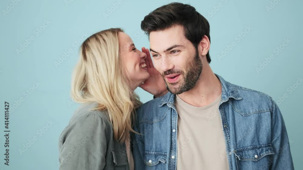 Emotional couple man and woman are sharing secrets while standing isolated over blue background in studio Emotional couple man and woman are sharing secrets while standing isolated over blue background in studio