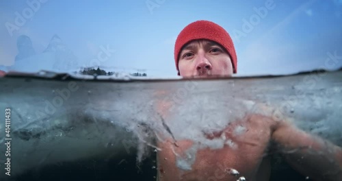 Winter swimmer portrait, cold treatment. Young man soaks in the icy winter lake then turns his head and looks at the camera. Split composition with underwater view photo