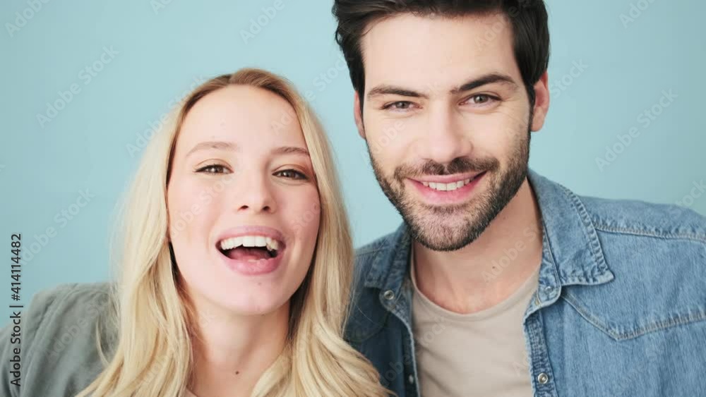 Playful couple man and woman are posing to the camera standing isolated over blue background in studio Playful couple man and woman are posing to the camera standing isolated over blue background in studio