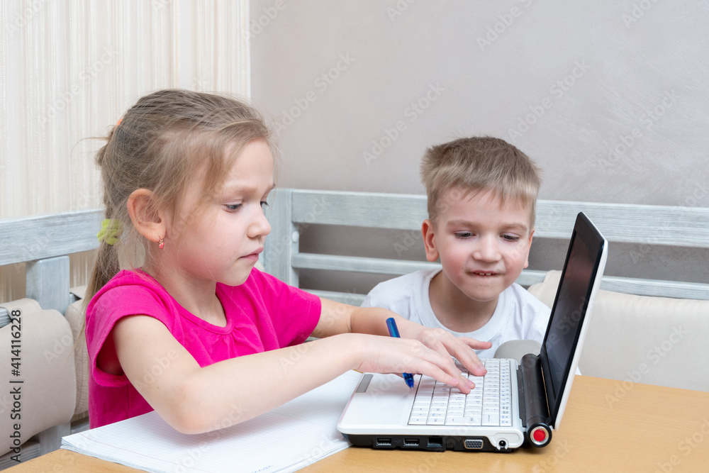 Little boy and girl study online at home. Cute little girl student shows how to do homework to her brother. The girl is typing the text on the laptop. Learning concept, distance learning