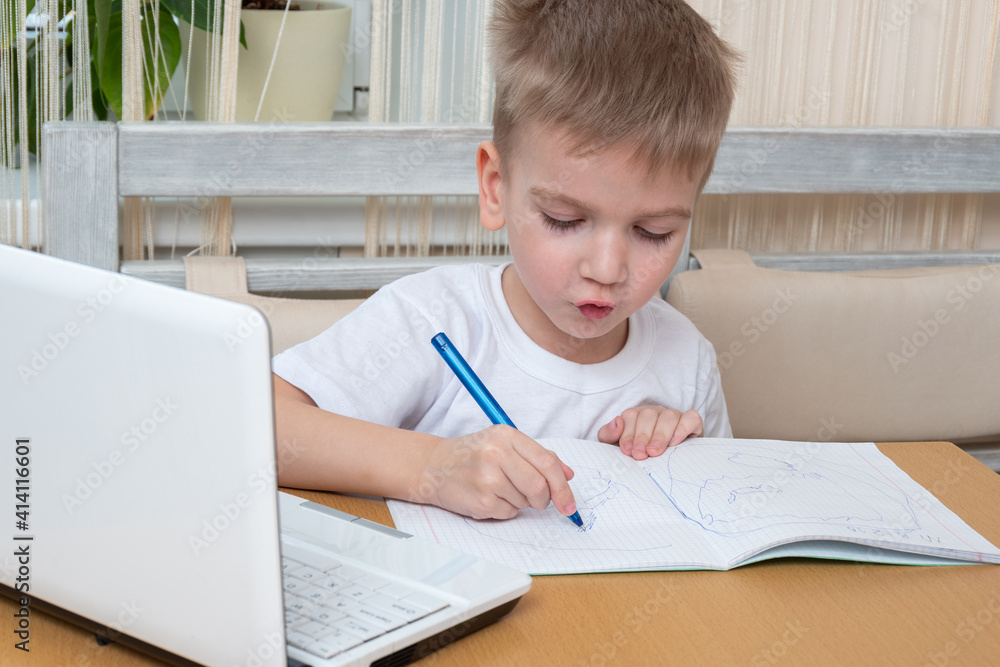The child studies online at home. Cute little boy draws homework in a notebook sitting at the table in front of a laptop. Online home education concept The child studies online at home. Cute little boy draws homework in a notebook sitting at the table in front of a laptop. Online home education concept