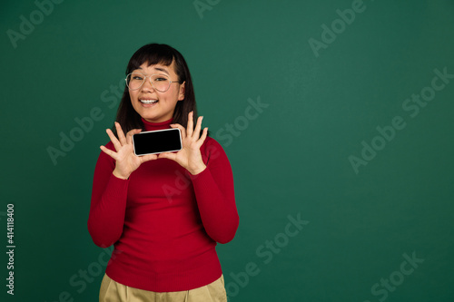 Shows blank phone screen. East asian young beautiful woman's portrait on green background with copyspace. Brunette female model. Concept of human emotions, facial expression, sales, ad, fashion. © master1305