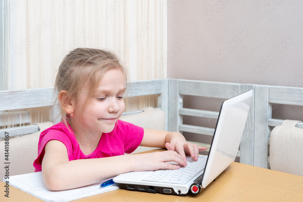 A cute little girl is sitting at a table and typing homework on a laptop at home. Learning concept, distance learning, self education, video conferencing lesson with mentor A cute little girl is sitting at a table and typing homework on a laptop at home. Learning concept, distance learning, self education, video conferencing lesson with mentor