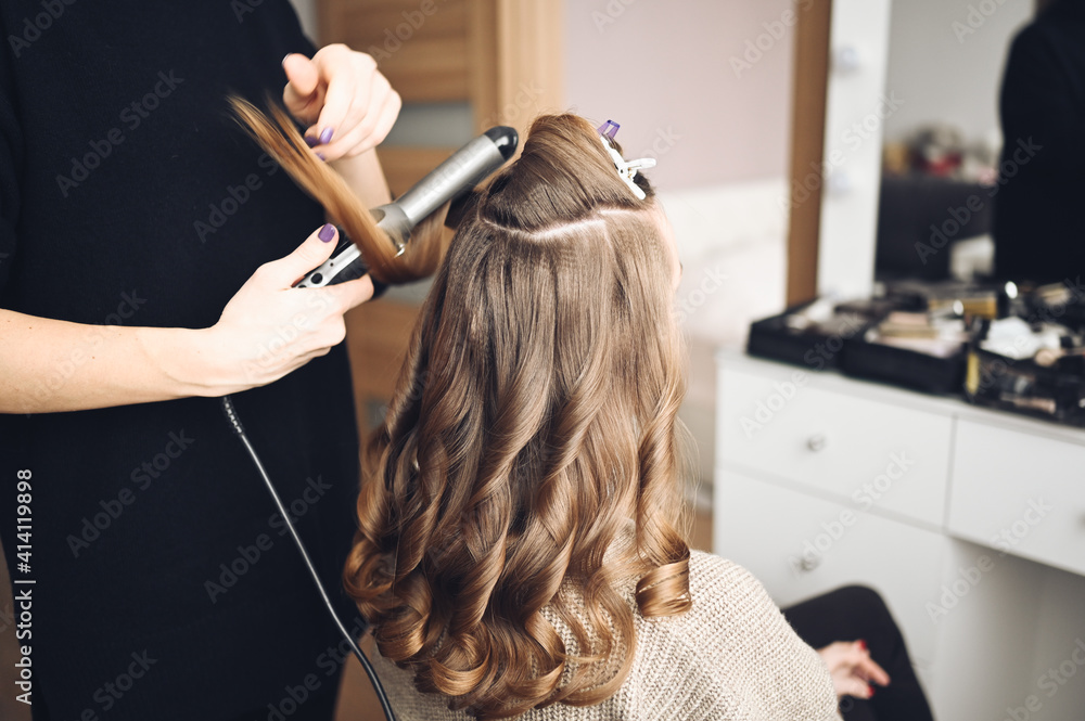Fototapeta premium Hair stylist prepares beautiful young woman for event, makes curls hairstyle with a curling iron for client in beauty salon. Long beautiful light brown natural hair.