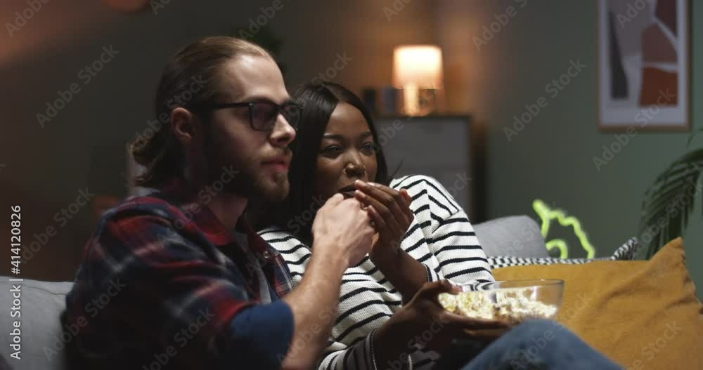 Close up of young multinational couple sitting at home on sofa and watching horror movie on tv at night. Beautiful girl frightened hides in the arms of guy.
