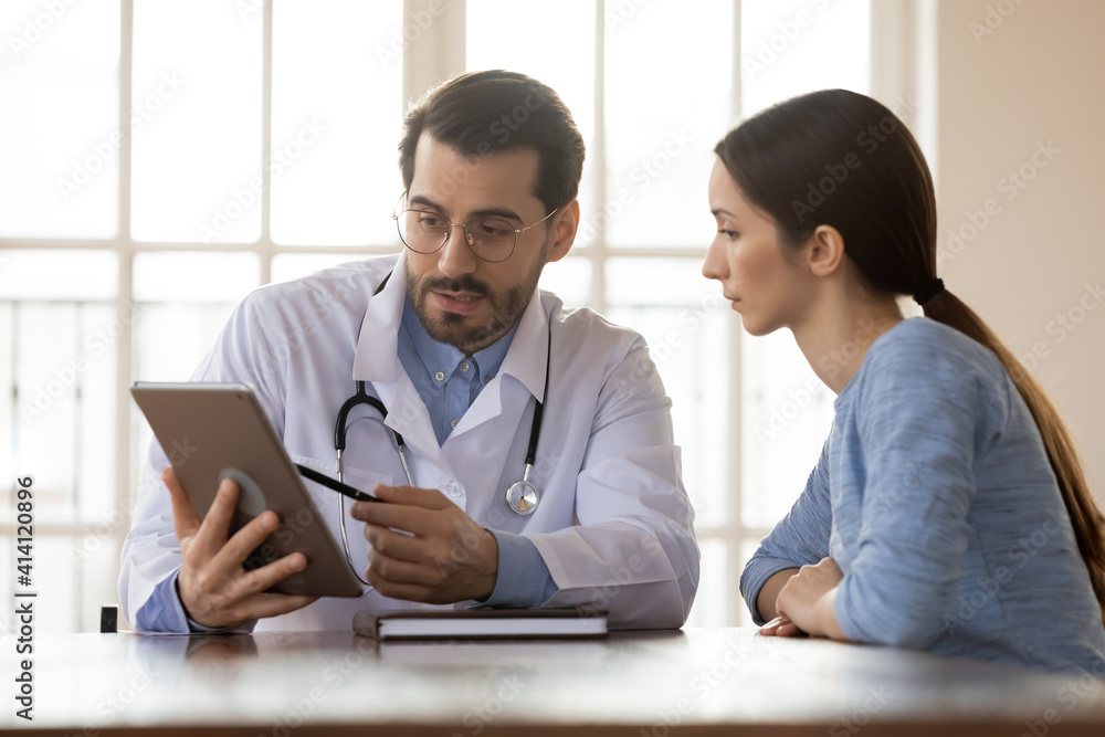 Focused young male doctor pointing at digital computer tablet screen, sharing health tests laboratory results to serious female patient or explaining medical insurance benefits at meeting in clinic. Focused young male doctor pointing at digital computer tablet screen, sharing health tests laboratory results to serious female patient or explaining medical insurance benefits at meeting in clinic.