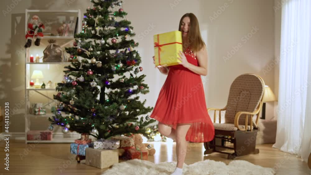 Wide shot portrait of happy excited pregnant woman dancing with Christmas gift at New Year tree at home. Young positive Caucasian lady enjoying holiday indoors. Fun and pregnancy concept. Wide shot portrait of happy excited pregnant woman dancing with Christmas gift at New Year tree at home. Young positive Caucasian lady enjoying holiday indoors. Fun and pregnancy concept.