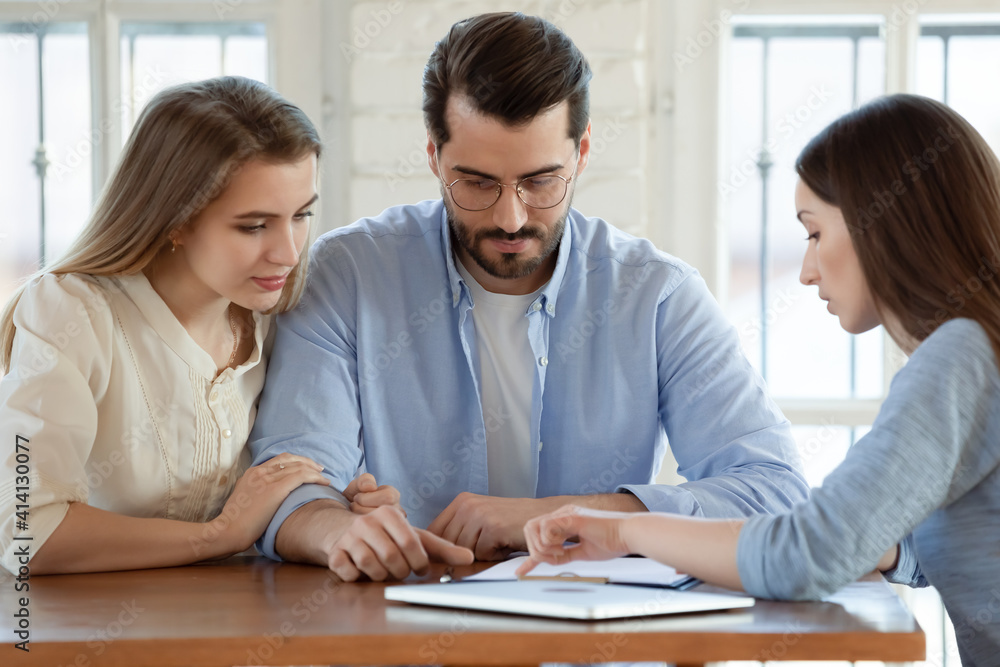 Focused young married couple discussing agreement with skilled real estate agent or broker. Professional financial advisor or saleswoman explaining contract details with interested clients at meeting. Focused young married couple discussing agreement with skilled real estate agent or broker. Professional financial advisor or saleswoman explaining contract details with interested clients at meeting.