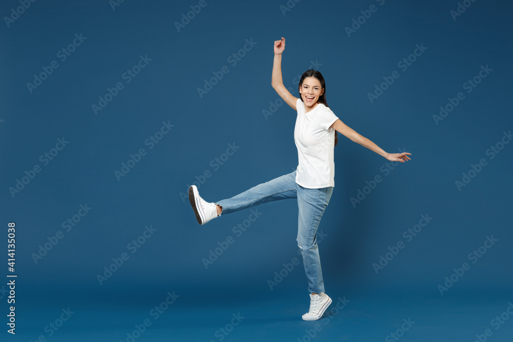Full length of young smiling fun relaxed beautiful latin woman 20s wearing white basic t-shirt keep hands outstretched with raised up leg look camera isolated on dark blue background studio portrait. Full length of young smiling fun relaxed beautiful latin woman 20s wearing white basic t-shirt keep hands outstretched with raised up leg look camera isolated on dark blue background studio portrait.