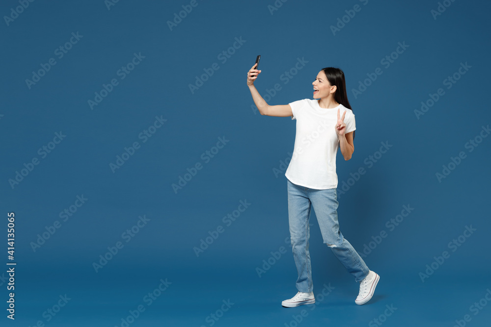 Full length side view of young happy friendly cheerful latin woman 20s in white t-shirt doing selfie shot on mobile phone show victory v-sign gesture isolated on dark blue background studio portrait.