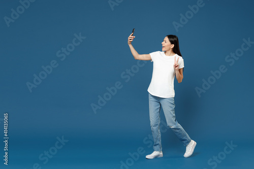 Full length side view of young happy friendly cheerful latin woman 20s in white t-shirt doing selfie shot on mobile phone show victory v-sign gesture isolated on dark blue background studio portrait.