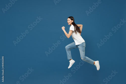 Full length side view of young happy sportive overjoyed brunette latin woman 20s wearing white casual basic t-shirt running fast jumping high isolated on dark blue color background studio portrait.