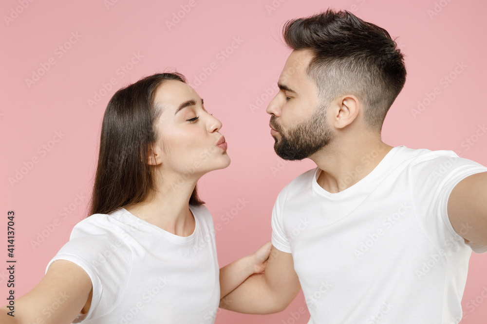 Close up young cheerful couple two friends man brunette woman in white basic blank print design t-shirt doing selfie shot on mobile cell phone isolated on pastel pink color background studio portrait Close up young cheerful couple two friends man brunette woman in white basic blank print design t-shirt doing selfie shot on mobile cell phone isolated on pastel pink color background studio portrait