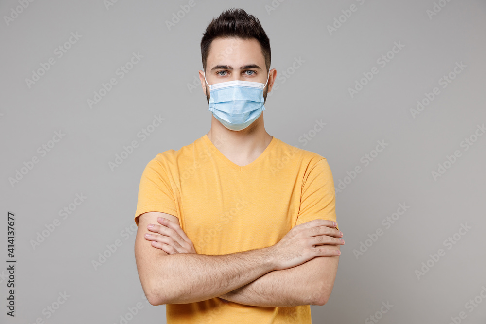 Young caucasian man 20s in yellow basic t-shirt in sterile face mask to safe from coronavirus virus covid-19 pandemic quarantine hold hands crossed folded isolated on grey background studio portrait. Young caucasian man 20s in yellow basic t-shirt in sterile face mask to safe from coronavirus virus covid-19 pandemic quarantine hold hands crossed folded isolated on grey background studio portrait.