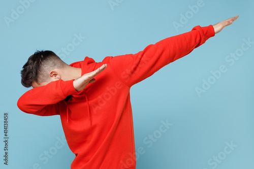 Young caucasian man 20s in casual red orange hoodie doing dab hip hop dance hands move gesture youth sign hiding covering face isolated on blue background studio portrait People lifestyle concept. photo