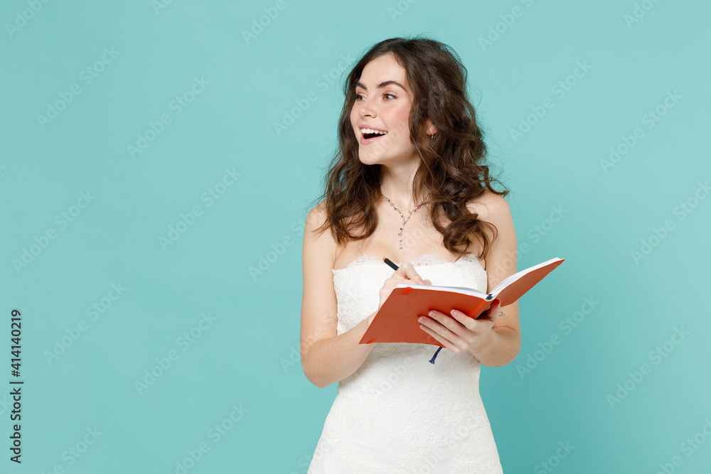 Cheerful bride young woman 20s years old wearing beautiful white wedding dress writing notes on notebook looking aside isolated on blue turquoise color background. Ceremony celebration party concept. Cheerful bride young woman 20s years old wearing beautiful white wedding dress writing notes on notebook looking aside isolated on blue turquoise color background. Ceremony celebration party concept.