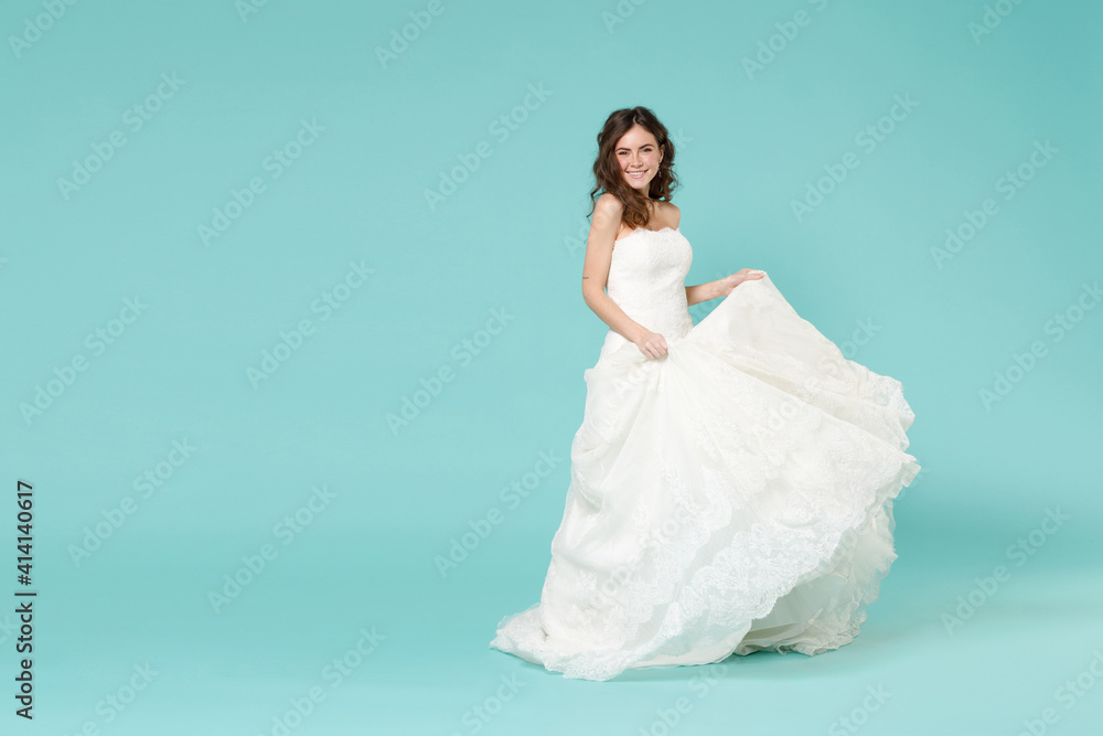 Full length of smiling tender bride woman 20s in beautiful white wedding dress standing looking camera isolated on blue turquoise color background studio portrait. Ceremony celebration party concept. Full length of smiling tender bride woman 20s in beautiful white wedding dress standing looking camera isolated on blue turquoise color background studio portrait. Ceremony celebration party concept.