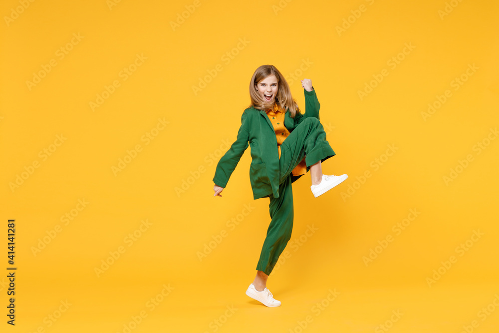 Full length little blonde smiling kid girl 12-13 years old in casual clothes do winner gesture clench fist raised up leg isolated on yellow background children portrait. Childhood lifestyle concept