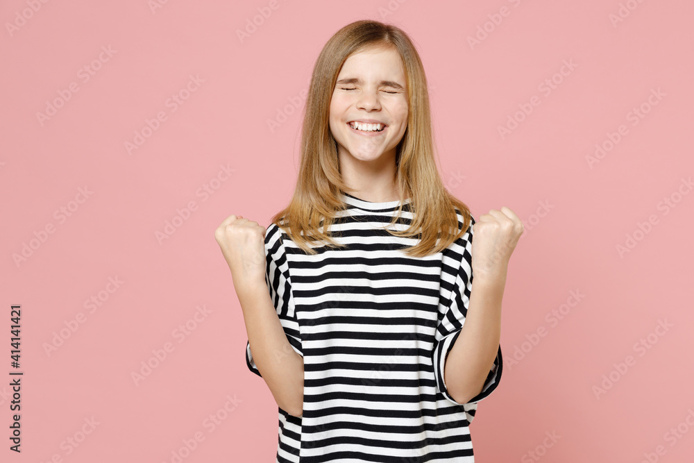 Little blonde cute kid girl 12-13 years old wearing striped oversized t-shirt do winner gesture clench fist isolated on pastel pink background children studio portrait. Childhood lifestyle concept. Little blonde cute kid girl 12-13 years old wearing striped oversized t-shirt do winner gesture clench fist isolated on pastel pink background children studio portrait. Childhood lifestyle concept.