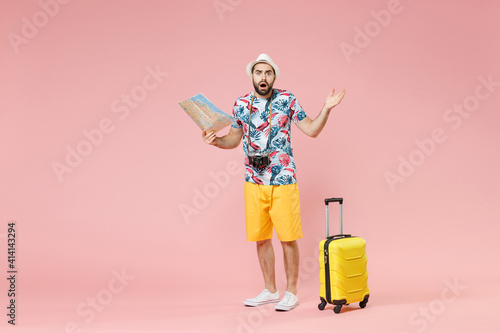 Full length of worried shocked young traveler tourist man in summer clothes hat hold city map spreading hands isolated on pink background. Passenger traveling on weekends. Air flight journey concept.