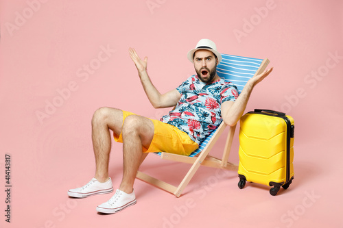 Foto Full length of shocked worried young traveler tourist man in summer clothes hat sit on deck chair spreading hands isolated on pink background