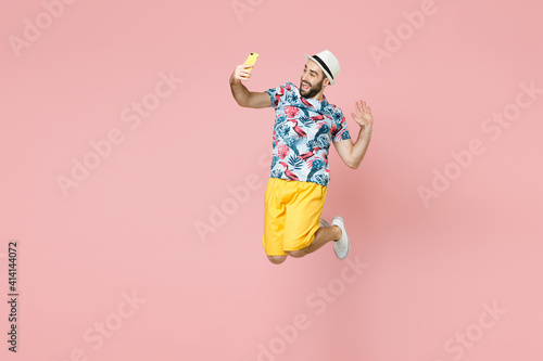 Full length of cheerful traveler tourist man in summer clothes hat jumping doing selfie shot on mobile phone isolated on pink background. Passenger traveling on weekend. Air flight journey concept.