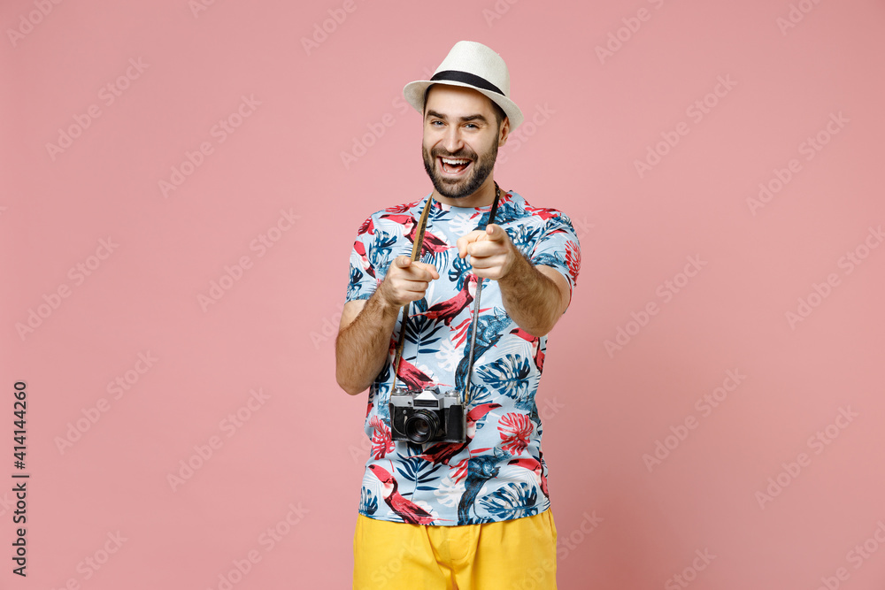 Cheerful young traveler tourist man in hat photo camera pointing index fingers camera on you isolated on pink background. Passenger traveling abroad on weekends getaway. Air flight journey concept.
